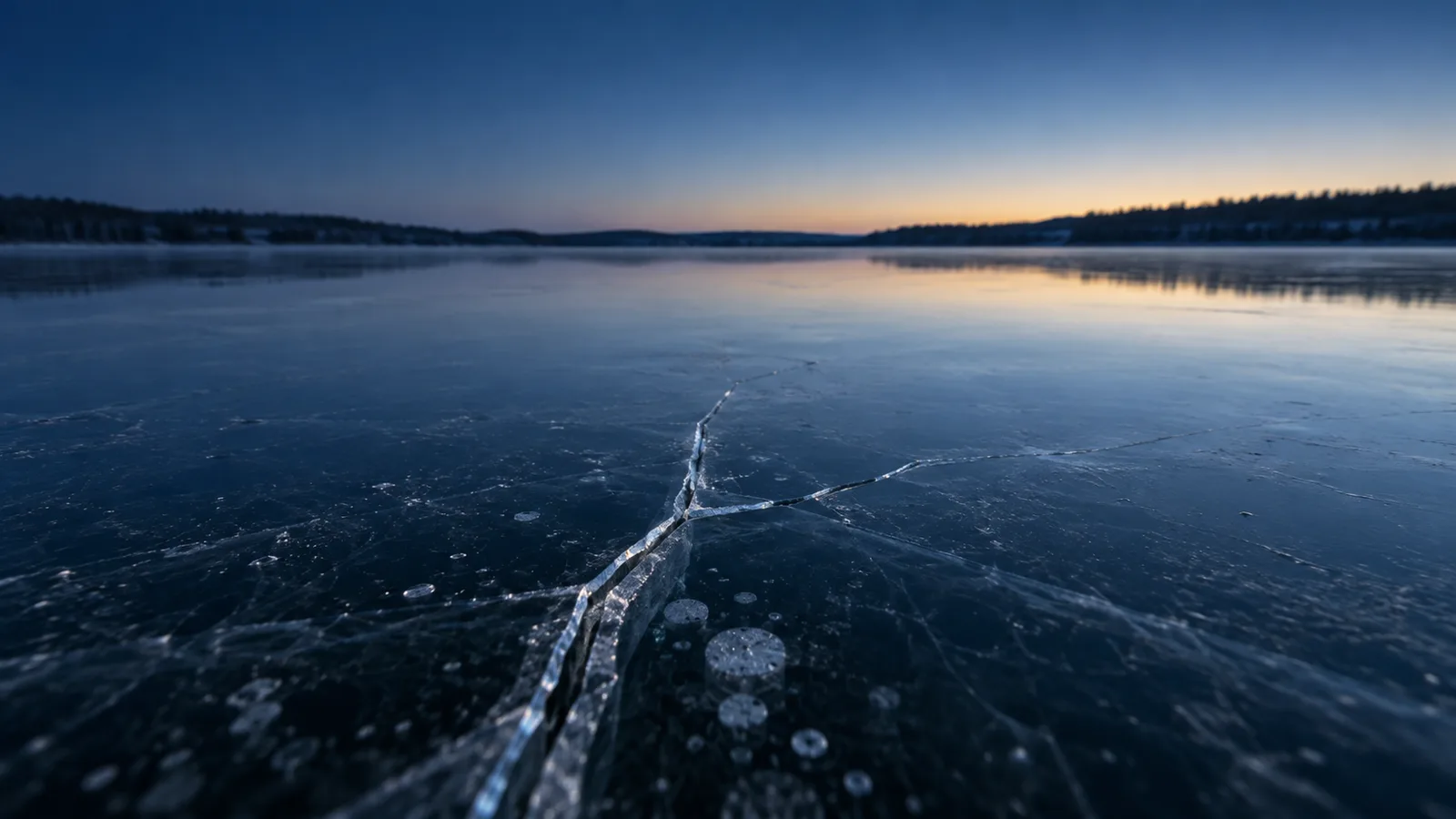 Superficie de hielo oscuro surcada de fisuras finas, con una grieta grande y ramificada en primer plano y burbujas atrapadas bajo el hielo, y el atardecer en el horizonte: imagen de la tesis de Hyman Minsky de que la estabilidad prolongada incuba la fragilidad financiera